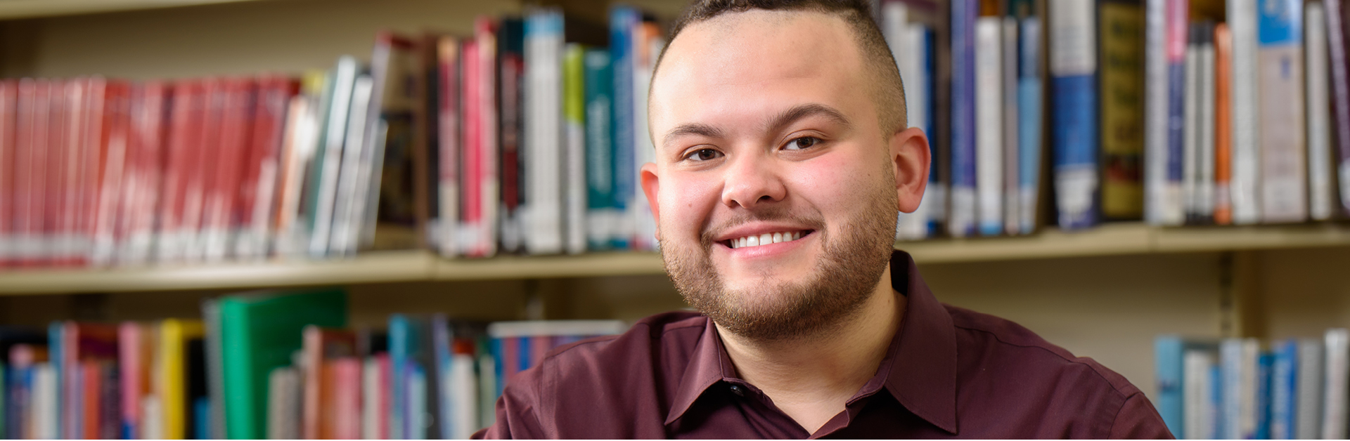 Photo of a man sitting in front of a laptop with a wall of bookshelves behind him. 