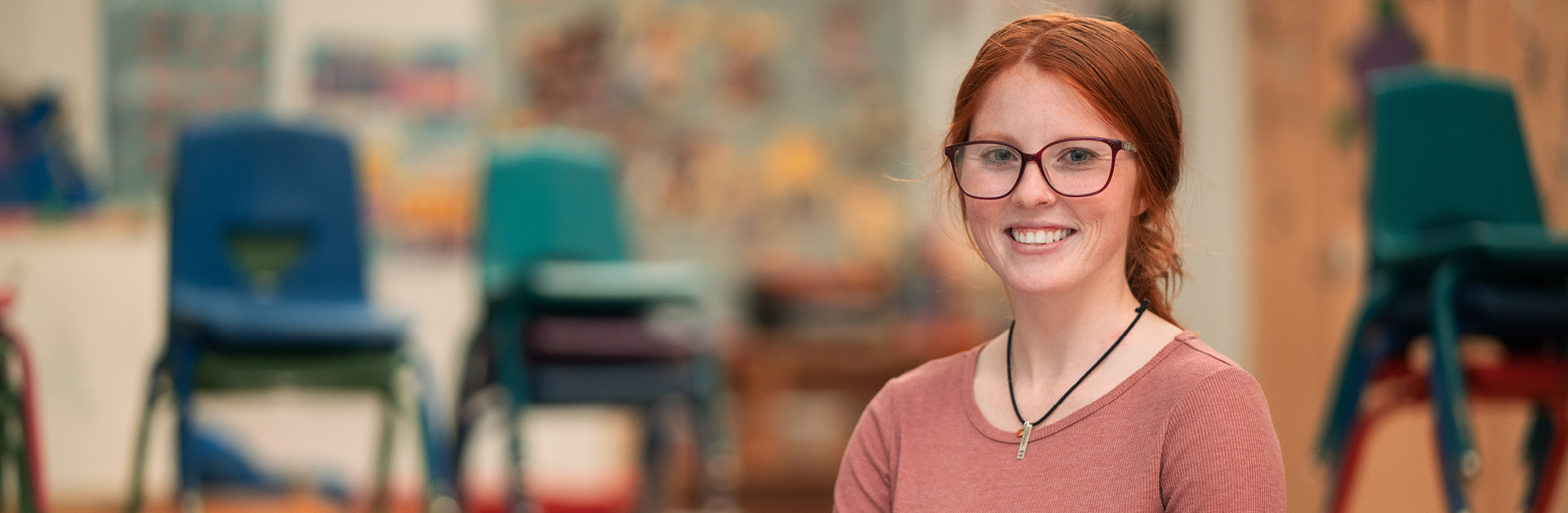 A female teacher with red hair and glasses stands in a classroom smiling