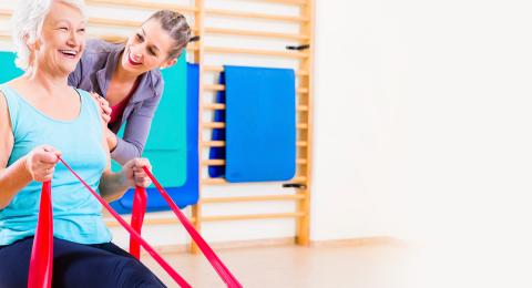 An older person in a gym working with resistance bands while a younger person leans in to talk to them