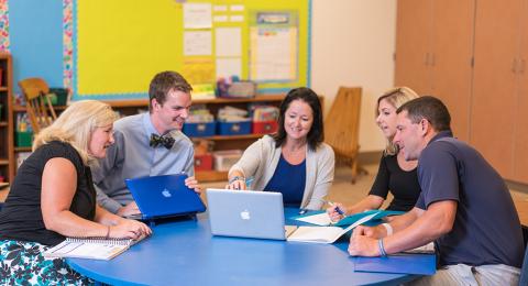 Five educators around a round classroom table sharing information on their laptops