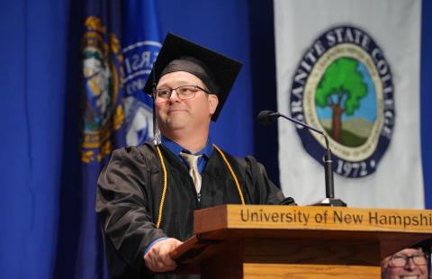 Photo of a man wearing graduation gown and hat, standing at a podium delivering a graduation speech