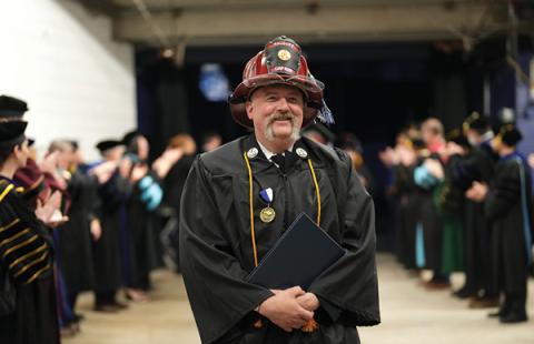 photo of a man wearing a fireman hat and graduation gown at a commencement ceremony