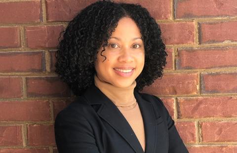 A headshot of a woman wearing a blazer standing in front of a brick wall