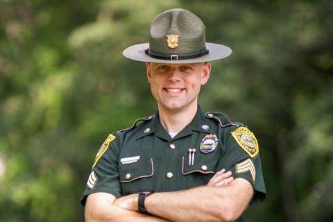 Male state trooper smiling and standing in front of trees with arms crossed