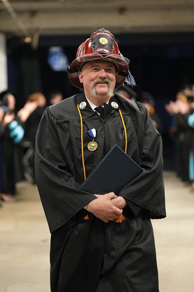 photo of a man wearing fireman hat and graduation gown at commencement ceremony