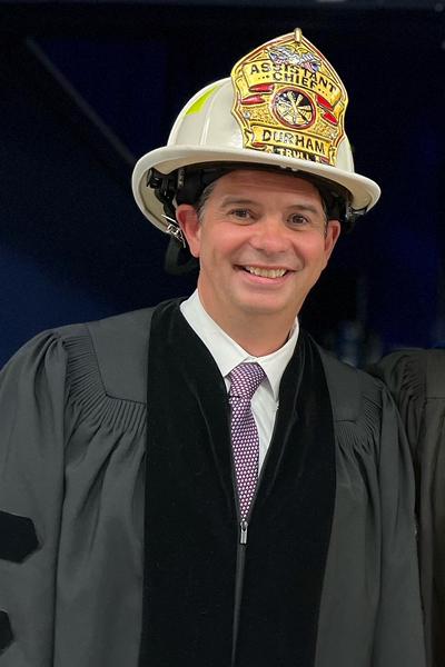 photo of a man wearing a fireman hat and faculty gown at graduation ceremony