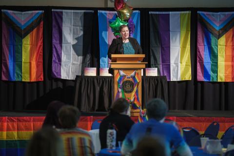 LGBTQ event woman at podium with rainbow flags behind her
